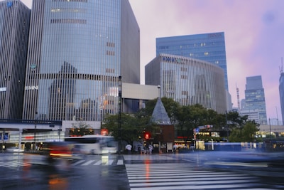 A bustling urban scene featuring tall modern buildings with glass facades. The street is busy with people and vehicles, creating a dynamic and lively atmosphere. Trees and a small building with a pointed roof provide contrast amidst the towering structures.