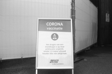 A black and white photograph features a signboard standing on a pavement. The sign is related to a COVID-19 vaccination site, with text in Dutch advising visitors to wear a mask due to the inability to always maintain a distance of 1.5 meters. The background includes a tent or temporary structure.
