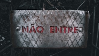 A weathered metal sign with a diamond pattern and rusty patches displays the text 'NÃO ENTRE' in red letters. It is affixed with bolts at both ends, set against a dark, blurred background of foliage and metal structures.