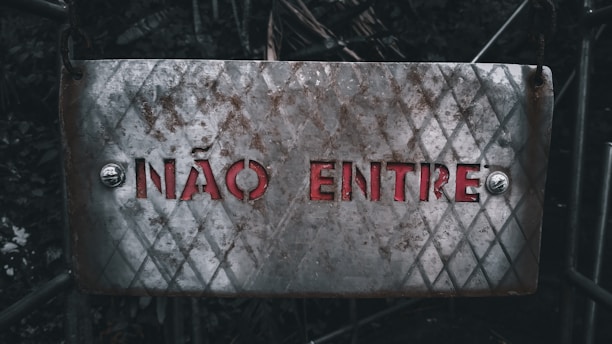 A weathered metal sign with a diamond pattern and rusty patches displays the text 'NÃO ENTRE' in red letters. It is affixed with bolts at both ends, set against a dark, blurred background of foliage and metal structures.