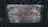 A weathered metal sign with a diamond pattern and rusty patches displays the text 'NÃO ENTRE' in red letters. It is affixed with bolts at both ends, set against a dark, blurred background of foliage and metal structures.