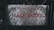 A weathered metal sign with a diamond pattern and rusty patches displays the text 'NÃO ENTRE' in red letters. It is affixed with bolts at both ends, set against a dark, blurred background of foliage and metal structures.