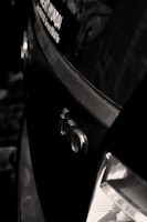 A close-up shot of the rear section of a car with an Audi logo visible, along with part of a dealership sticker on the window. The tail light is partially visible and the car appears to be black, with reflections suggesting a glossy finish.