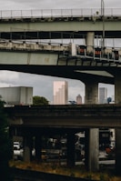 A complex network of elevated highways and overpasses frames a city skyline in the background. Several tall buildings are visible beyond the highways, with one distinctively brown skyscraper standing out. The scene has an urban, industrial feel with visible infrastructure details like traffic signs and heavy vehicles.