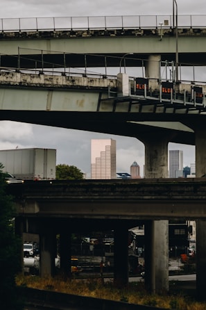 A complex network of elevated highways and overpasses frames a city skyline in the background. Several tall buildings are visible beyond the highways, with one distinctively brown skyscraper standing out. The scene has an urban, industrial feel with visible infrastructure details like traffic signs and heavy vehicles.
