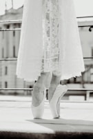 A black and white image of a ballet dancer standing on pointe with a delicate lace skirt reaching just above the ankles. The focus is on the dancer's feet, highlighting the elegance and control of the pose. The background appears to be an urban setting with blurred architectural elements.