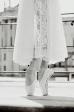 A black and white image of a ballet dancer standing on pointe with a delicate lace skirt reaching just above the ankles. The focus is on the dancer's feet, highlighting the elegance and control of the pose. The background appears to be an urban setting with blurred architectural elements.