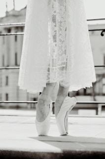 A black and white image of a ballet dancer standing on pointe with a delicate lace skirt reaching just above the ankles. The focus is on the dancer's feet, highlighting the elegance and control of the pose. The background appears to be an urban setting with blurred architectural elements.