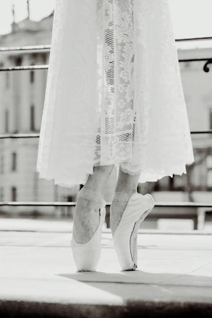 A black and white image of a ballet dancer standing on pointe with a delicate lace skirt reaching just above the ankles. The focus is on the dancer's feet, highlighting the elegance and control of the pose. The background appears to be an urban setting with blurred architectural elements.
