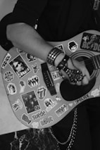 A person playing an acoustic guitar adorned with numerous colorful stickers, including band logos and artistic designs. The person's hand is wearing a studded glove and a bracelet, adding an edgy style to the scene.