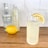 A tall glass of a lemon-flavored beverage garnished with a lemon slice sits on a wooden surface. Behind the glass, there is a lemon, a bottle of Tanqueray Sterling imported gin, and a bottle of Sparkling Ice classic lemonade. The background features white tiled walls.