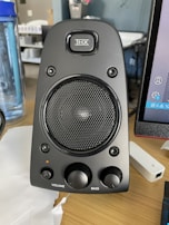 A black computer speaker equipped with a mesh-covered central driver and a brand logo above it. Two control knobs for volume and bass adjustment are located at the bottom. The surrounding area includes a blue water bottle on the left and part of a computer monitor on the right.