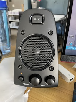 A black computer speaker equipped with a mesh-covered central driver and a brand logo above it. Two control knobs for volume and bass adjustment are located at the bottom. The surrounding area includes a blue water bottle on the left and part of a computer monitor on the right.