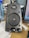 A black computer speaker equipped with a mesh-covered central driver and a brand logo above it. Two control knobs for volume and bass adjustment are located at the bottom. The surrounding area includes a blue water bottle on the left and part of a computer monitor on the right.