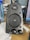 A black computer speaker equipped with a mesh-covered central driver and a brand logo above it. Two control knobs for volume and bass adjustment are located at the bottom. The surrounding area includes a blue water bottle on the left and part of a computer monitor on the right.