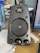 A black computer speaker equipped with a mesh-covered central driver and a brand logo above it. Two control knobs for volume and bass adjustment are located at the bottom. The surrounding area includes a blue water bottle on the left and part of a computer monitor on the right.