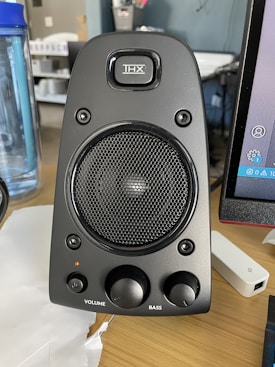 A black computer speaker equipped with a mesh-covered central driver and a brand logo above it. Two control knobs for volume and bass adjustment are located at the bottom. The surrounding area includes a blue water bottle on the left and part of a computer monitor on the right.
