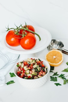 A white bowl filled with a colorful salad containing chopped cucumbers, tomatoes, red onions, and herbs is placed on a white surface. Nearby, three ripe red tomatoes with green stems rest on a white plate. A halved lemon and scattered parsley leaves add a touch of freshness, alongside a metal lemon squeezer.