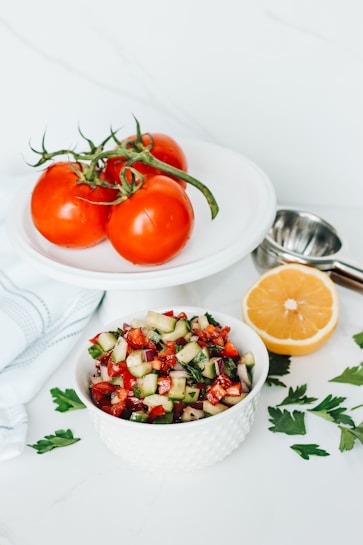 A white bowl filled with a colorful salad containing chopped cucumbers, tomatoes, red onions, and herbs is placed on a white surface. Nearby, three ripe red tomatoes with green stems rest on a white plate. A halved lemon and scattered parsley leaves add a touch of freshness, alongside a metal lemon squeezer.