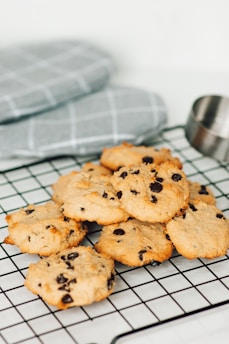 A pile of freshly baked chocolate chip cookies is arranged on a black cooling rack. In the background, there are grey and white checkered oven mitts and a stainless steel measuring cup.