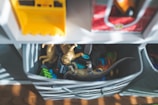 A storage unit holds bins filled with children's toys including dinosaur figures and toy vehicles. Sunlight filters through slats, casting shadows across the toys. The toys are in a soft fabric container, and shelves with more bins are visible above.