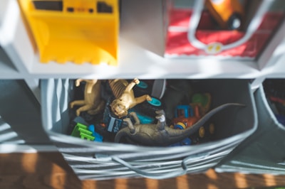 A storage unit holds bins filled with children's toys including dinosaur figures and toy vehicles. Sunlight filters through slats, casting shadows across the toys. The toys are in a soft fabric container, and shelves with more bins are visible above.