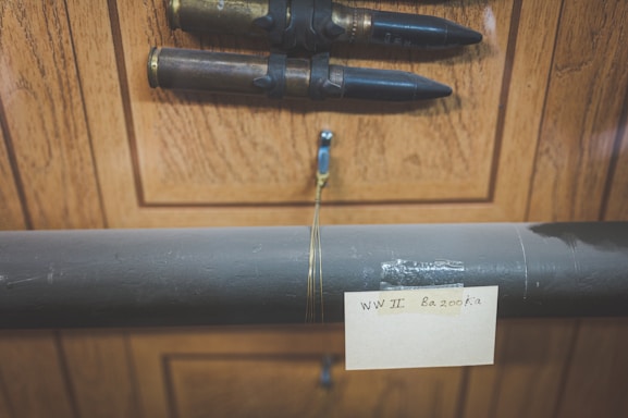 A wooden wall displays a World War II bazooka tube with a note attached, and two large bullets mounted above it.