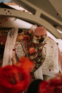 A couple is dressed in traditional wedding attire, adorned with intricate patterns and detailed embroidery. The bride wears a veil and jewelry, while the groom dons a turban with a feather. Flower petals are scattered in the air, creating a festive atmosphere.
