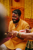 A man is joyfully smiling, wearing a traditional yellow attire, with red powder smeared on his face during a festive celebration. He is surrounded by colorful marigold garlands in the background, contributing to the vibrant and lively atmosphere. Hands with colorful bangles and henna designs hold a bowl of red powder in the foreground.