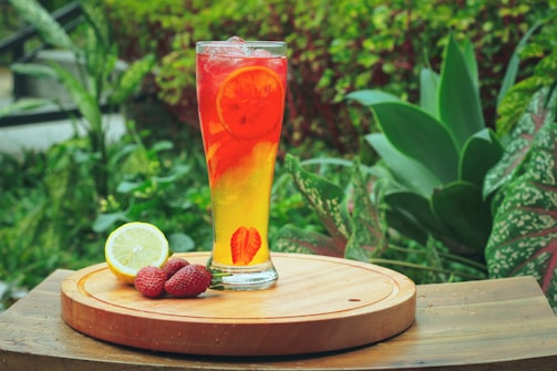 A tall glass filled with a vibrant, colorful drink containing slices of lemon and possibly strawberries, sits on a wooden surface. Beside the glass are a slice of lemon and some strawberries. The background features lush greenery, including various types of plants with large leaves.