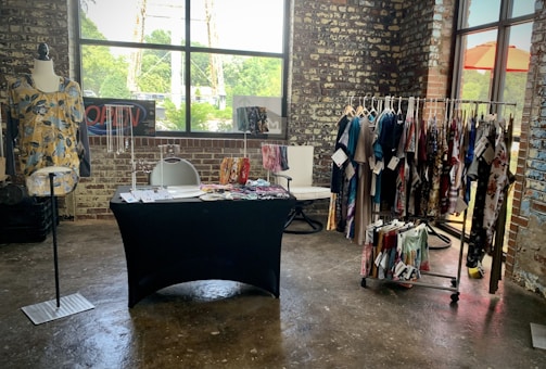 A rustic brick-walled room houses a small clothing display. A mannequin showcases a floral yellow top next to a table covered in a black cloth with accessories and compartments for organizing items. Behind the table, there is a chair with a modern design. A clothing rack neatly holds various colorful garments, while a window lets in natural light, highlighting the textures and colors in the space.