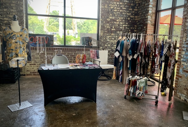 A rustic brick-walled room houses a small clothing display. A mannequin showcases a floral yellow top next to a table covered in a black cloth with accessories and compartments for organizing items. Behind the table, there is a chair with a modern design. A clothing rack neatly holds various colorful garments, while a window lets in natural light, highlighting the textures and colors in the space.