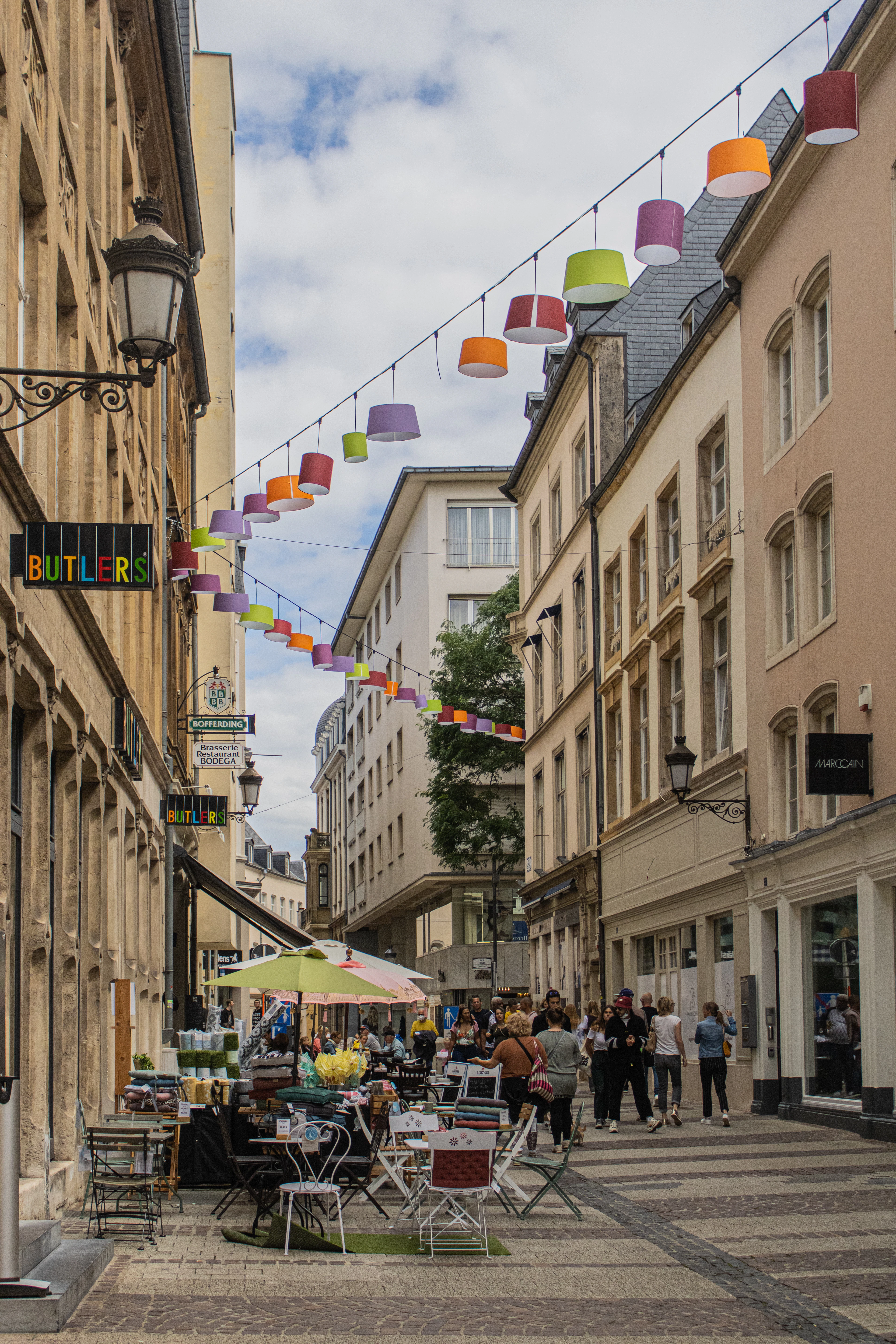 A pedestrian street in Luxembourg City's old town strung with rows of coloured lampshades above cafe terraces and shopfronts