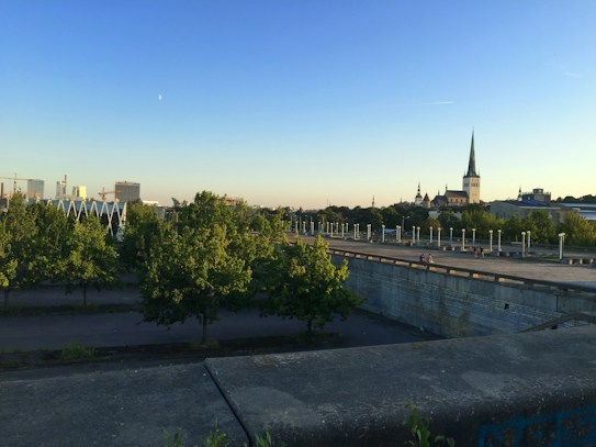 The image features a scenic urban landscape with a clear sky and visible crescent moon. A large open area is surrounded by numerous trees, creating a natural foreground. In the background, there are several buildings, including a prominent structure with a tall spire. The overall setting is peaceful and bathed in warm evening light.
