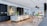 A modern kitchen with sleek, minimalistic design elements, featuring a large marble countertop island with seating, dark cabinetry, and contemporary light fixtures. There are plants and decor items on the island, adding a touch of greenery. The open living area includes a dining table with chairs and a cozy seating space, creating an inviting and functional atmosphere.