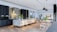 A modern kitchen with sleek, minimalistic design elements, featuring a large marble countertop island with seating, dark cabinetry, and contemporary light fixtures. There are plants and decor items on the island, adding a touch of greenery. The open living area includes a dining table with chairs and a cozy seating space, creating an inviting and functional atmosphere.