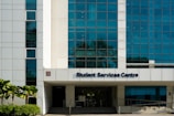 A modern building facade with large blue-tinted windows and a white exterior. The entrance is labeled Student Services Centre. Palms and shrubs are visible in the foreground, adding greenery.