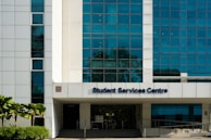 A modern building facade with large blue-tinted windows and a white exterior. The entrance is labeled Student Services Centre. Palms and shrubs are visible in the foreground, adding greenery.