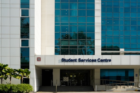 A modern building facade with large blue-tinted windows and a white exterior. The entrance is labeled Student Services Centre. Palms and shrubs are visible in the foreground, adding greenery.