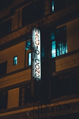 A multi-story building with illuminated vertical signage that reads 'Baguio Condo'. The facade is lit with a dim, moody light, highlighting the architectural features. Windows with a blue tint are visible, providing a nighttime urban atmosphere.