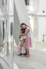 Two young girls stand together in a modern, minimalist setting with white walls and large geometric structures. One girl is leaning on another in a comforting embrace, both dressed in floral skirts and sandals.