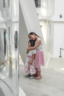 Two young girls stand together in a modern, minimalist setting with white walls and large geometric structures. One girl is leaning on another in a comforting embrace, both dressed in floral skirts and sandals.