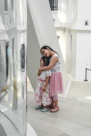 Two young girls stand together in a modern, minimalist setting with white walls and large geometric structures. One girl is leaning on another in a comforting embrace, both dressed in floral skirts and sandals.