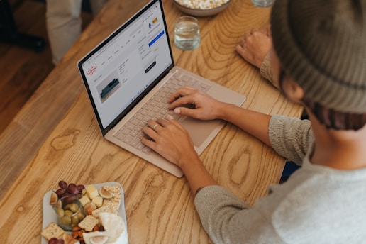 A person wearing a beanie and sweater is using a laptop on a wooden table. The screen displays an online shopping site. Next to the laptop, there are snacks including grapes, olives, crackers, and cheese on a plate. Another person is partially visible, sitting across the table, with a glass of water nearby.