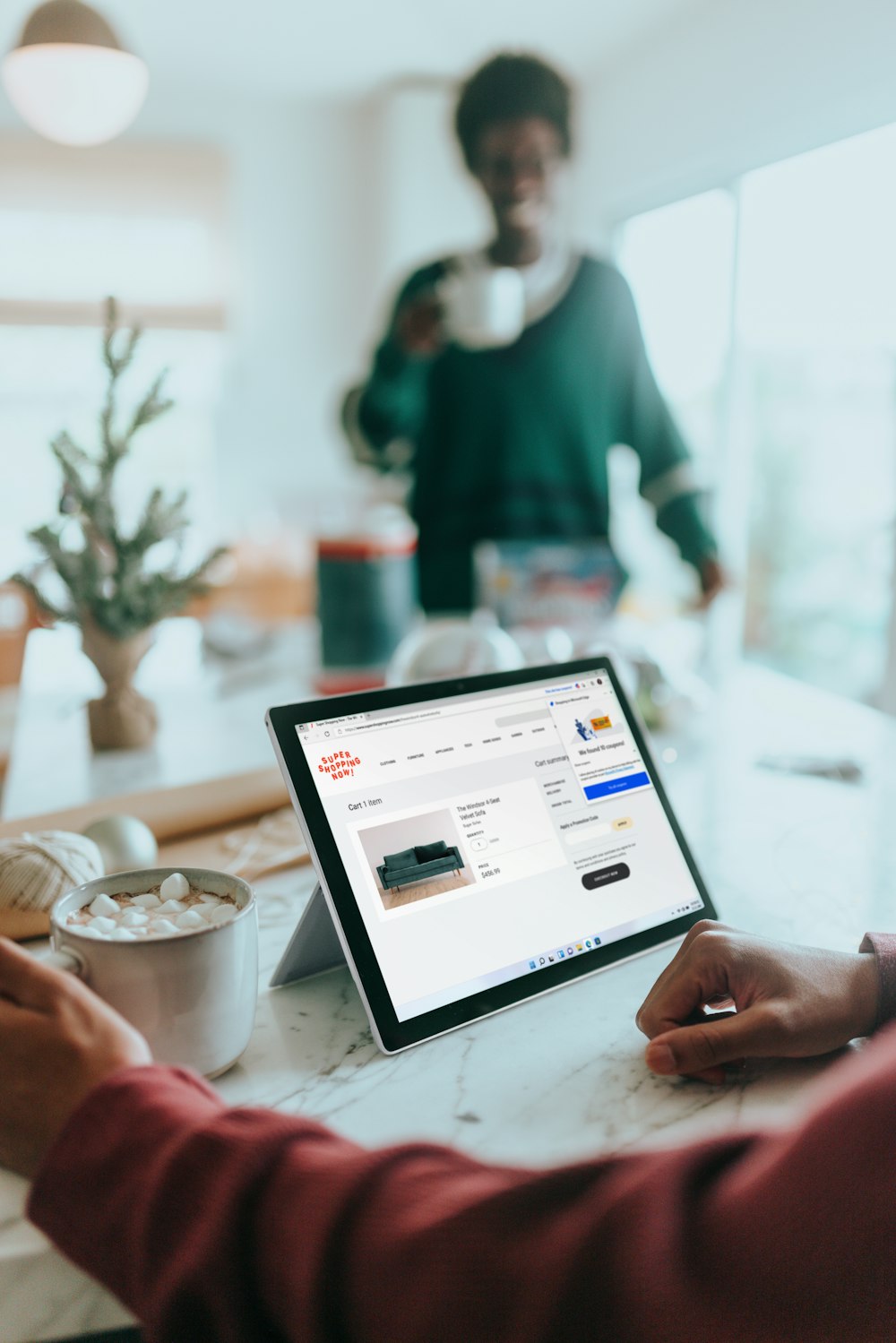 Person on their Surfacelaptop online shopping drinking hot chocolate with person in background out of focus