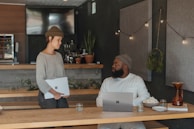 Two people are in a cozy indoor setting with a focus on working or conversing. One person is sitting at a wooden table with a laptop and a bowl of popcorn, while the other person, standing, holds what appears to be another laptop. The background includes decorative plants and string lights, contributing to a warm and inviting atmosphere.