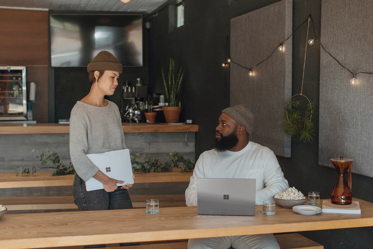 Two coworkers mid-conversation, one standing with a laptop handing work over to another seated with a laptop, the moment a task passes from one set of hands to the next.