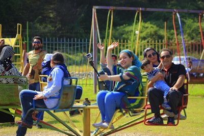 A group of people are enjoying a ride on a colorful playground merry-go-round. Among them, a woman with a bright smile holds a selfie stick, capturing their fun moment. In the background, more people are present, and a swing set and a fence surrounding the area are visible. The scene is set on a sunny day in a park or outdoor recreational area.