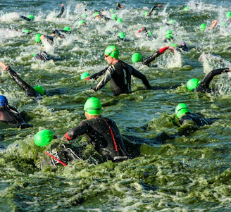 A large group of swimmers wearing green swim caps and wetsuits navigates through turbulent water, creating splashes as they make their way in a competitive swimming event.