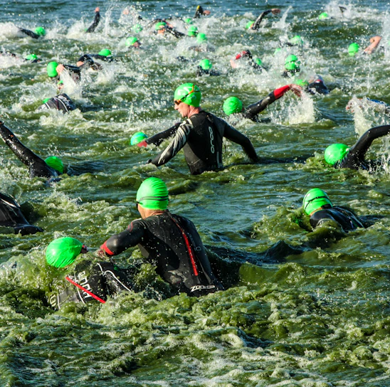 A large group of swimmers wearing green swim caps and wetsuits navigates through turbulent water, creating splashes as they make their way in a competitive swimming event.